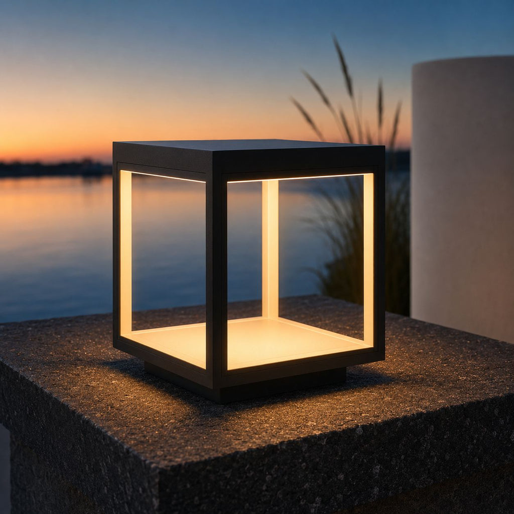 Warm white LED pillar light glowing on a granite plinth beside water at sunset