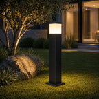 Slim black bollard light on a lawn beside an olive tree and boulder