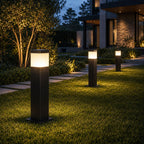 Three slim bollard lights along a planted path beside a modern stone house at dusk
