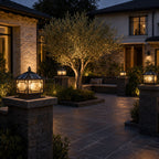 Two pillar lanterns on stone piers outside a stone house with an olive tree at dusk