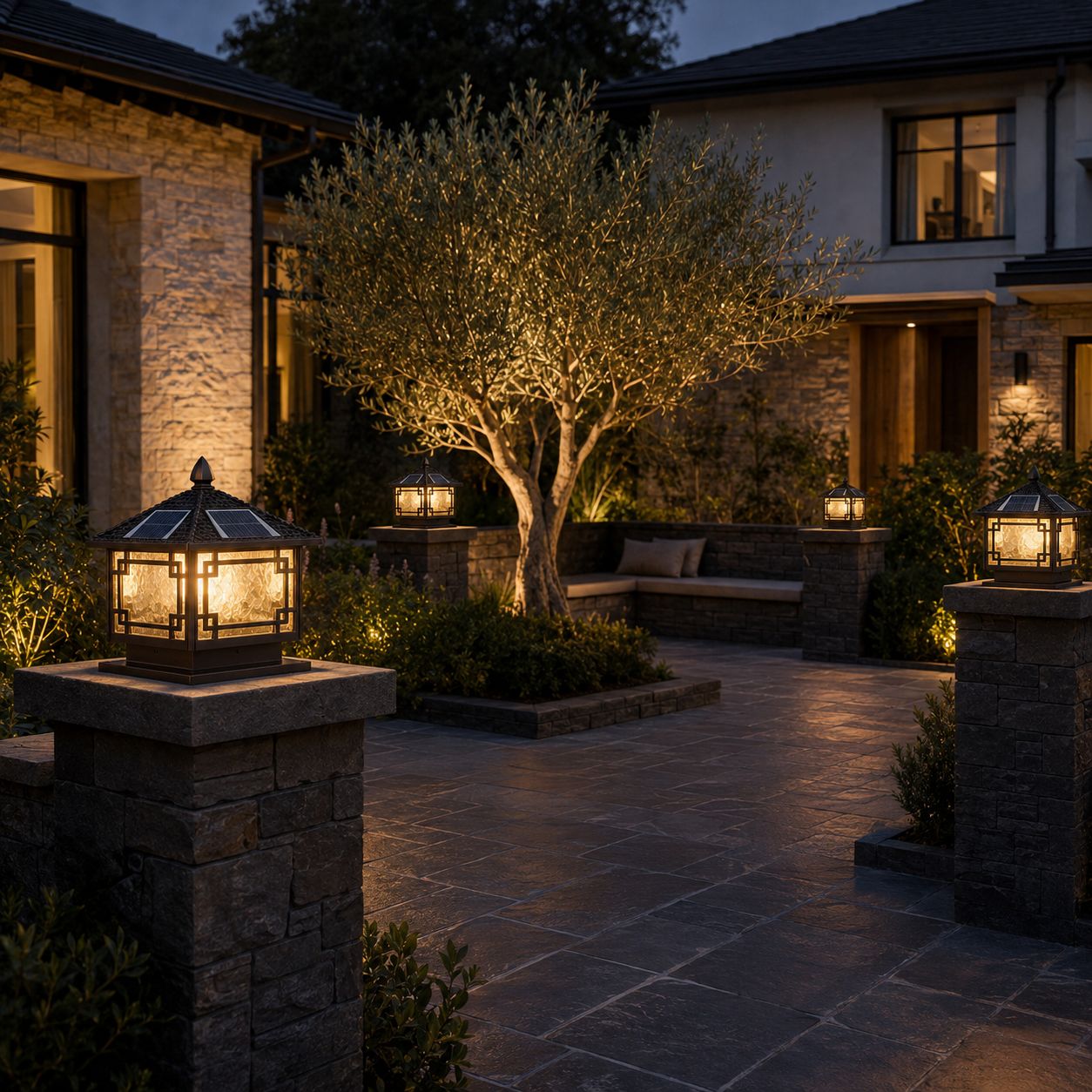 Two pillar lanterns on stone piers outside a stone house with an olive tree at dusk