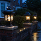 Three scrollwork pillar lights lit along a low brick wall on a wet street
