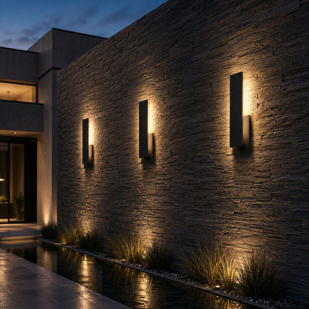 Three large slim wall lights on a dark stacked stone wall of a modern house at dusk