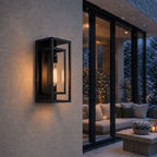 Outdoor wall sconce lit during snowfall beside a glass door and patio seating
