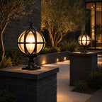 Two black globe post lights on charcoal brick walls at a modern entrance with olive trees