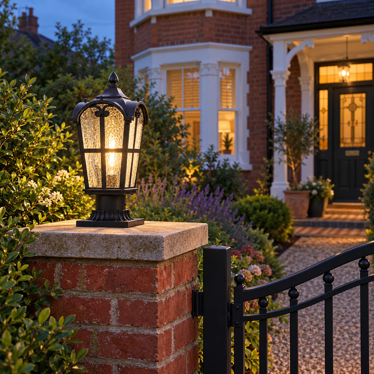Sentora black textured-glass pillar lantern illuminated on a brick gate pier beside a garden entrance at dusk.