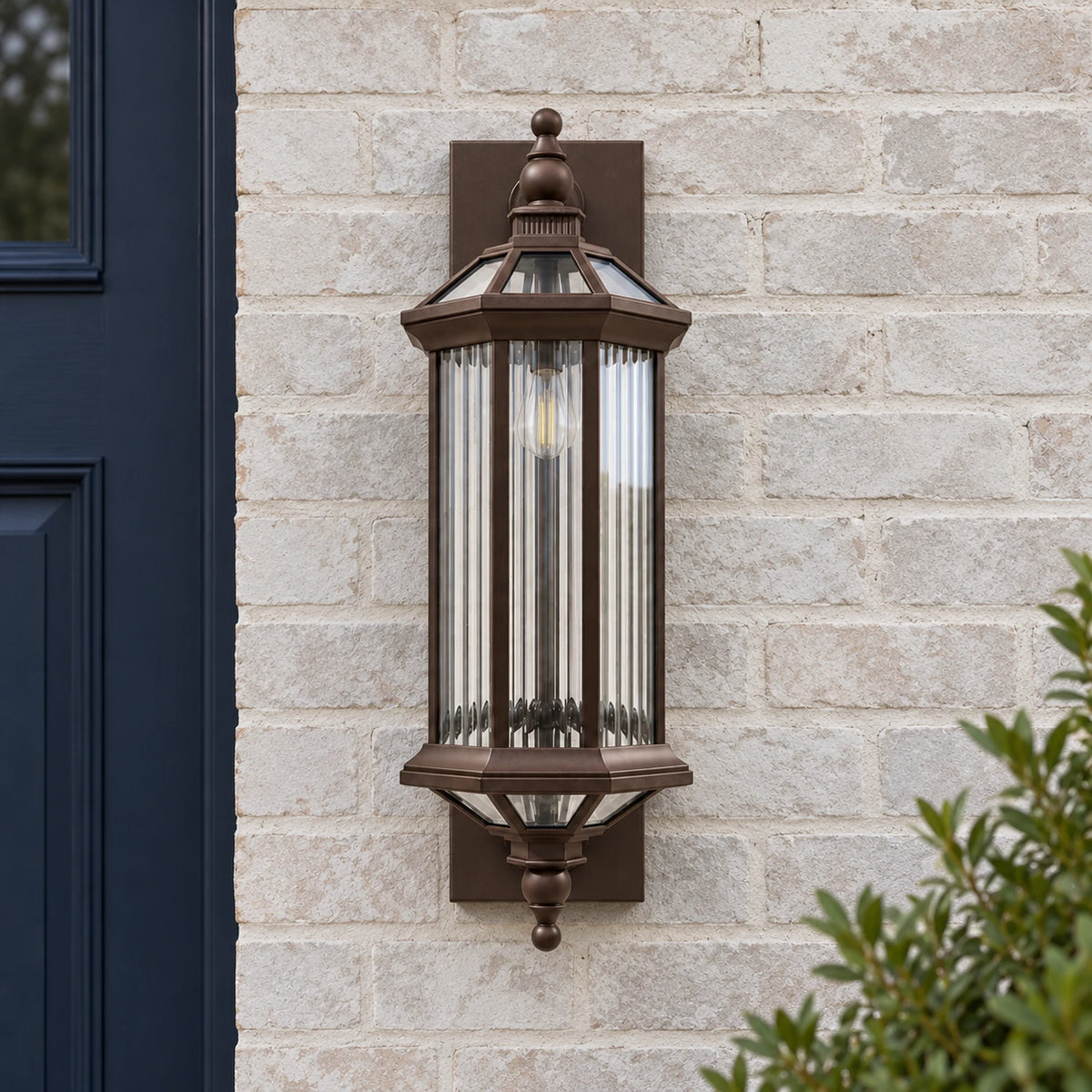 Unlit front view of the BronzeGlow bronze wall lantern with vertically textured clear glass on a pale brick wall.