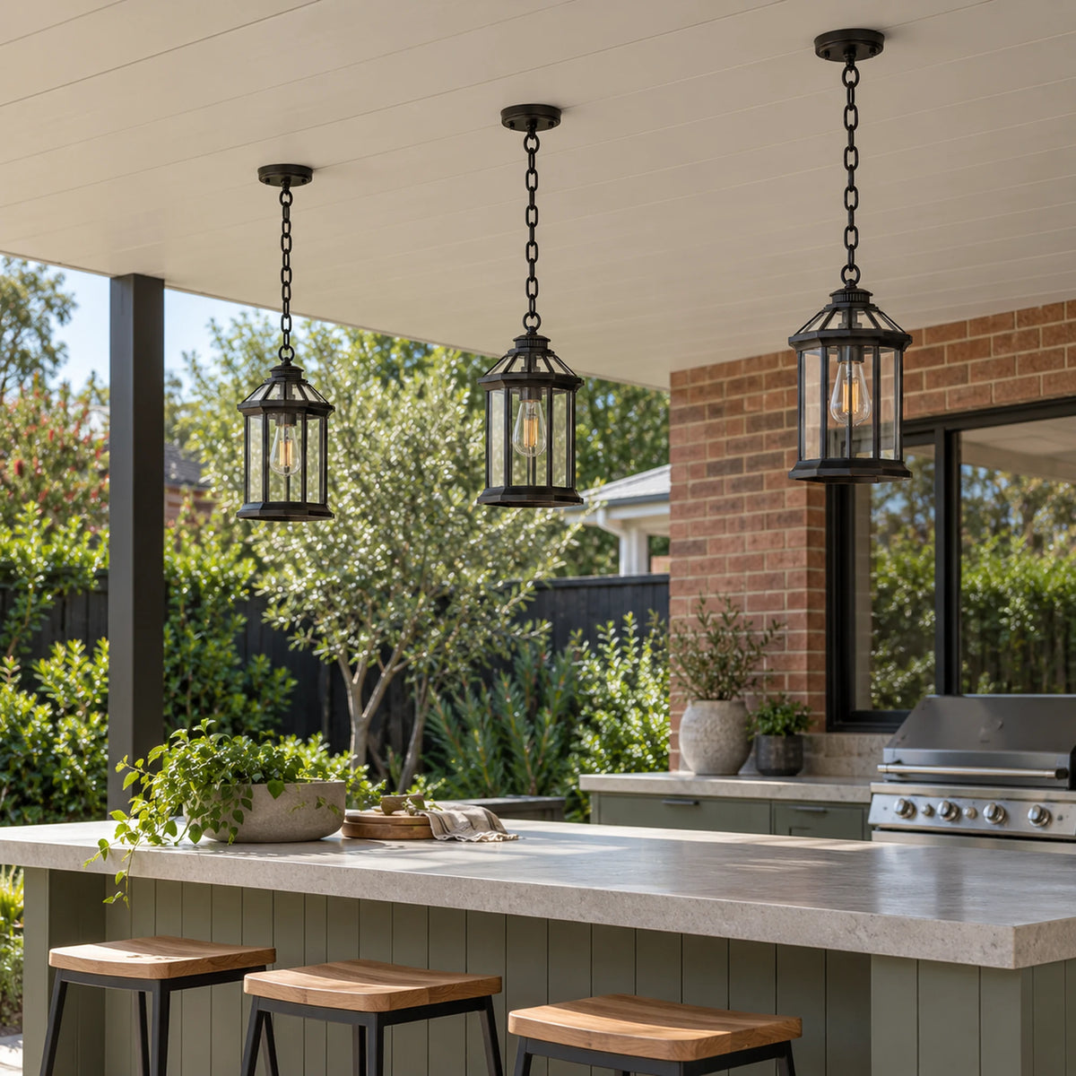 Three black Portiva glass pendants above a covered outdoor kitchen island with stools and a brick wall.